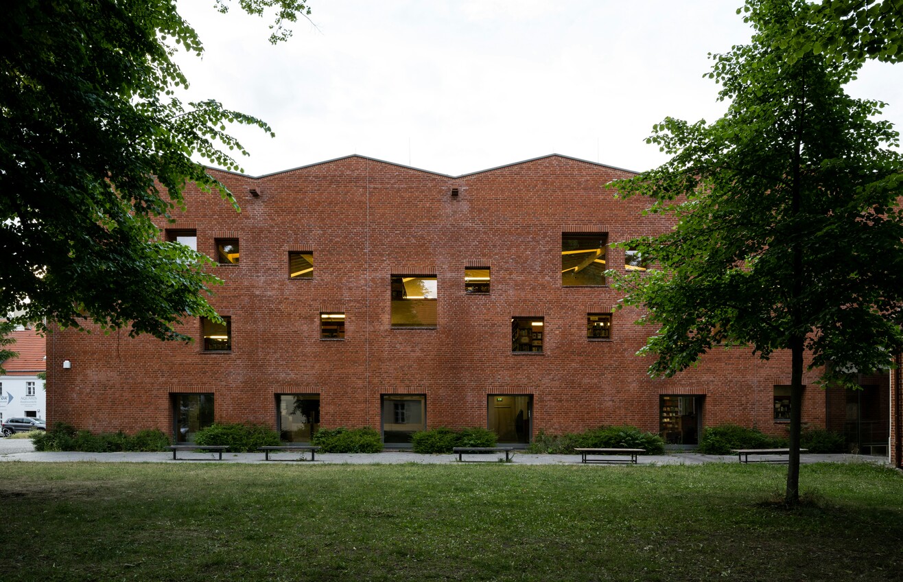 Backsteingebäude mit großen Fenstern, umgeben von grünen Bäumen und Sträuchern.