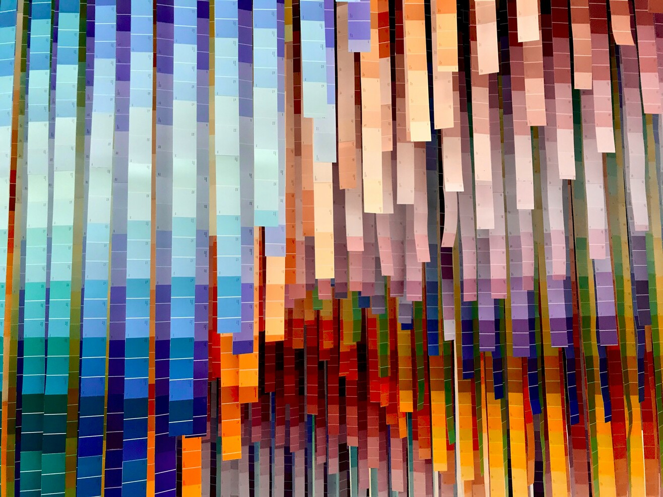 Abstrakte Wandinstallation aus vielen farbigen, vertikal hängenden Papierstreifen, die einen verlaufenden Regenbogeneffekt erzeugen.