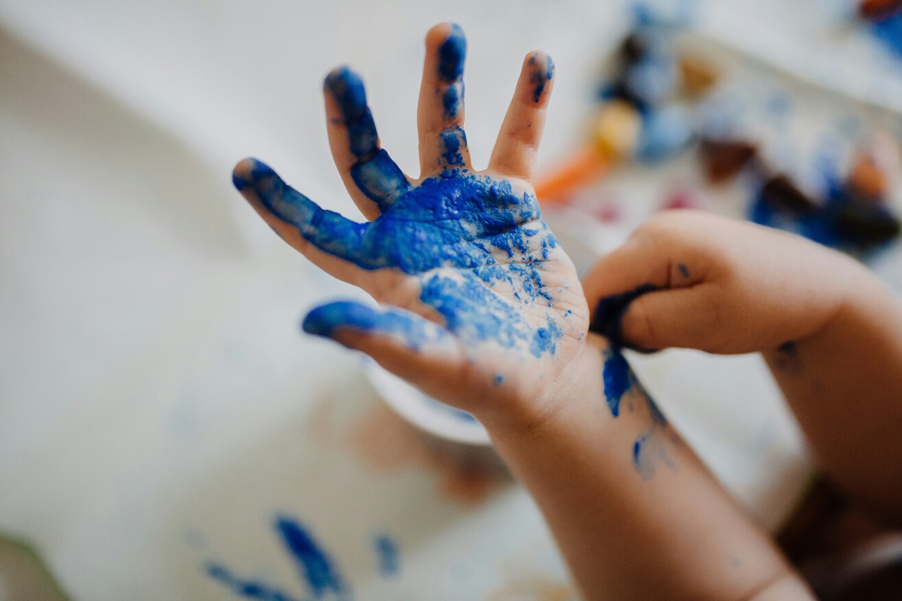 Eine Hand mit blauer Farbe vor einem bemalten Hintergrund beim kreativen Gestalten.