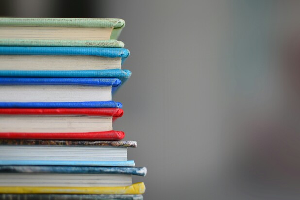 Nahaufnahme mehrerer übereinanderliegender Bücher mit unterschiedlich farbigen Einbänden. Der Hintergrund ist unscharf.