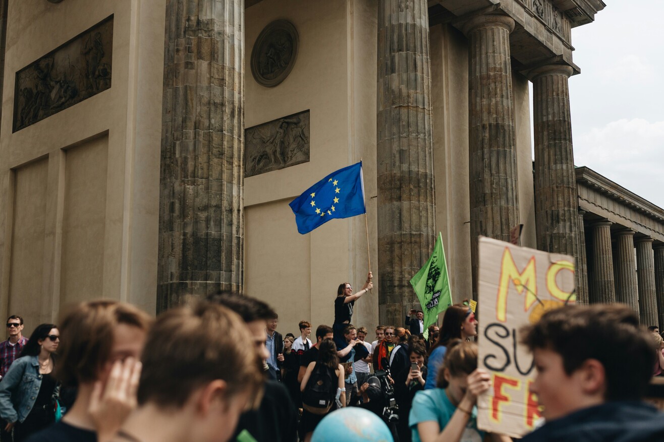Menschen bei einer Demonstration vor dem Brandenburger Tor in Berlin mit hochgehaltenen Schildern und Fahnen.