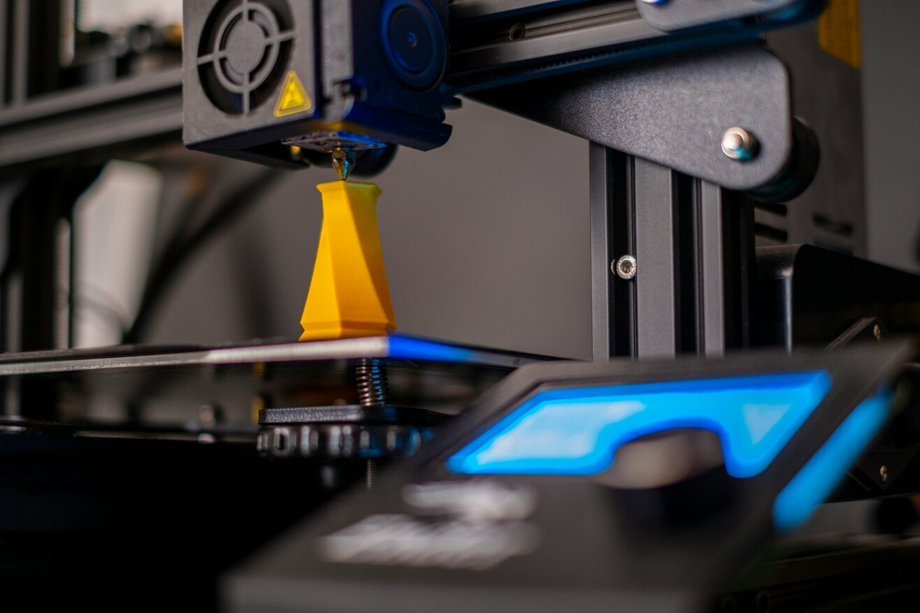 Nahaufnahme eines 3D-Druckers während des Druckvorgangs, dessen Arbeitsbereich von blauem Licht beleuchtet wird.