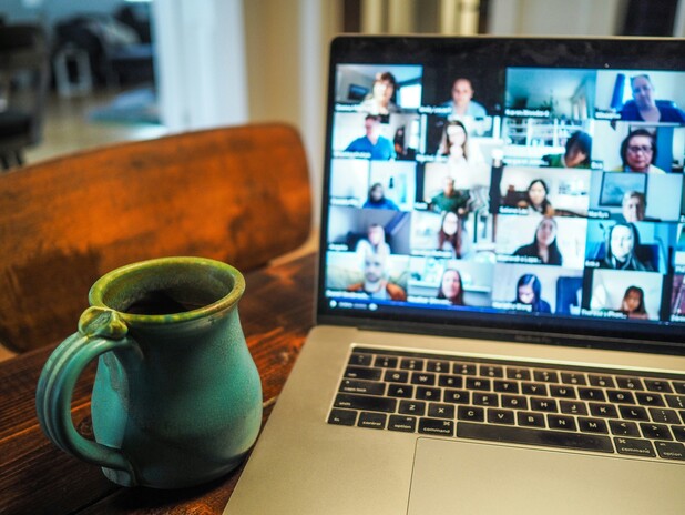 Eine geöffnete Laptopkamera zeigt eine Videokonferenz mit vielen Teilnehmenden in einem Rasterlayout. Daneben steht auf einem Holztisch eine grüne Keramiktasse mit Kaffee. Das Bild vermittelt eine typische Arbeitssituation im Homeoffice oder bei einem Online-Meeting.