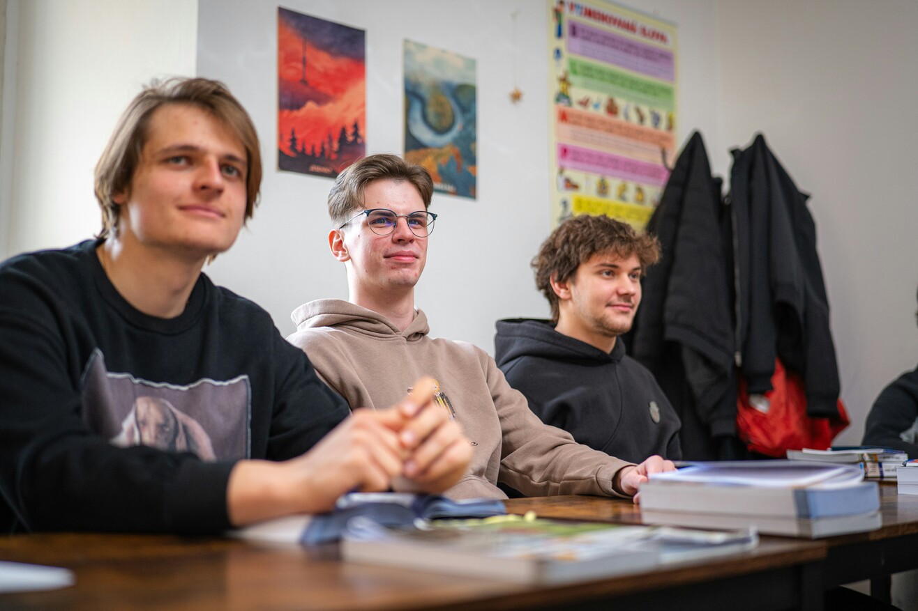 Drei junge Menschen sitzen an einem Tisch in einem hellen Klassen- oder Seminarraum. Sie blicken aufmerksam nach vorne und hören einer Person außerhalb des Bildes zu. Auf dem Tisch liegen Hefte und Schreibmaterialien. Im Hintergrund sind weitere Sitzplätze und der moderne Unterrichtsraum zu erkennen.
