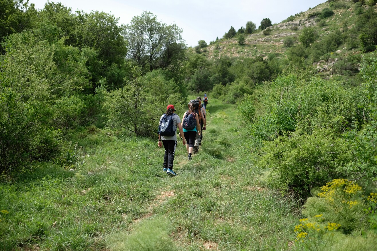 Eine Gruppe junger Menschen wandert mit Rucksäcken über eine grüne Wiese in einer weitläufigen Landschaft.