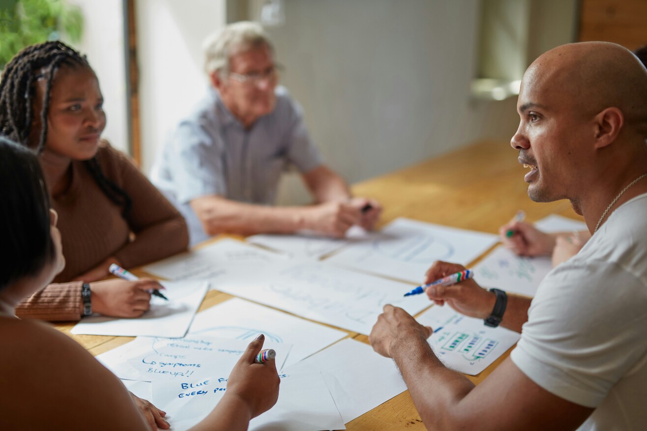 Mehrere Personen sitzen an einem Tisch und arbeiten gemeinsam mit Papieren und Stiften an einem Projekt.