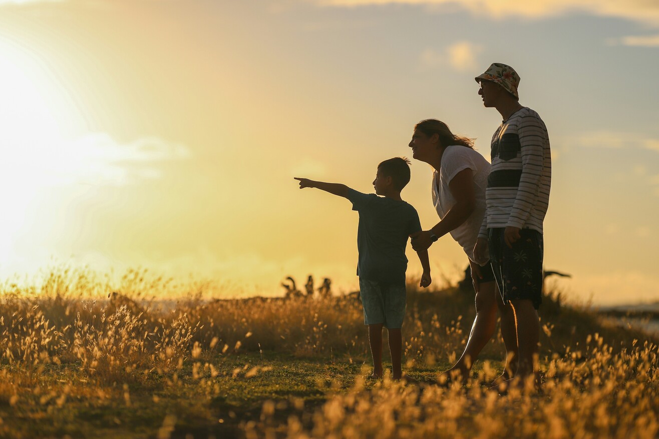 Eine Familie mit Mutter, Vater und Kind geht bei Sonnenuntergang über eine Wiese und hält sich an den Händen.