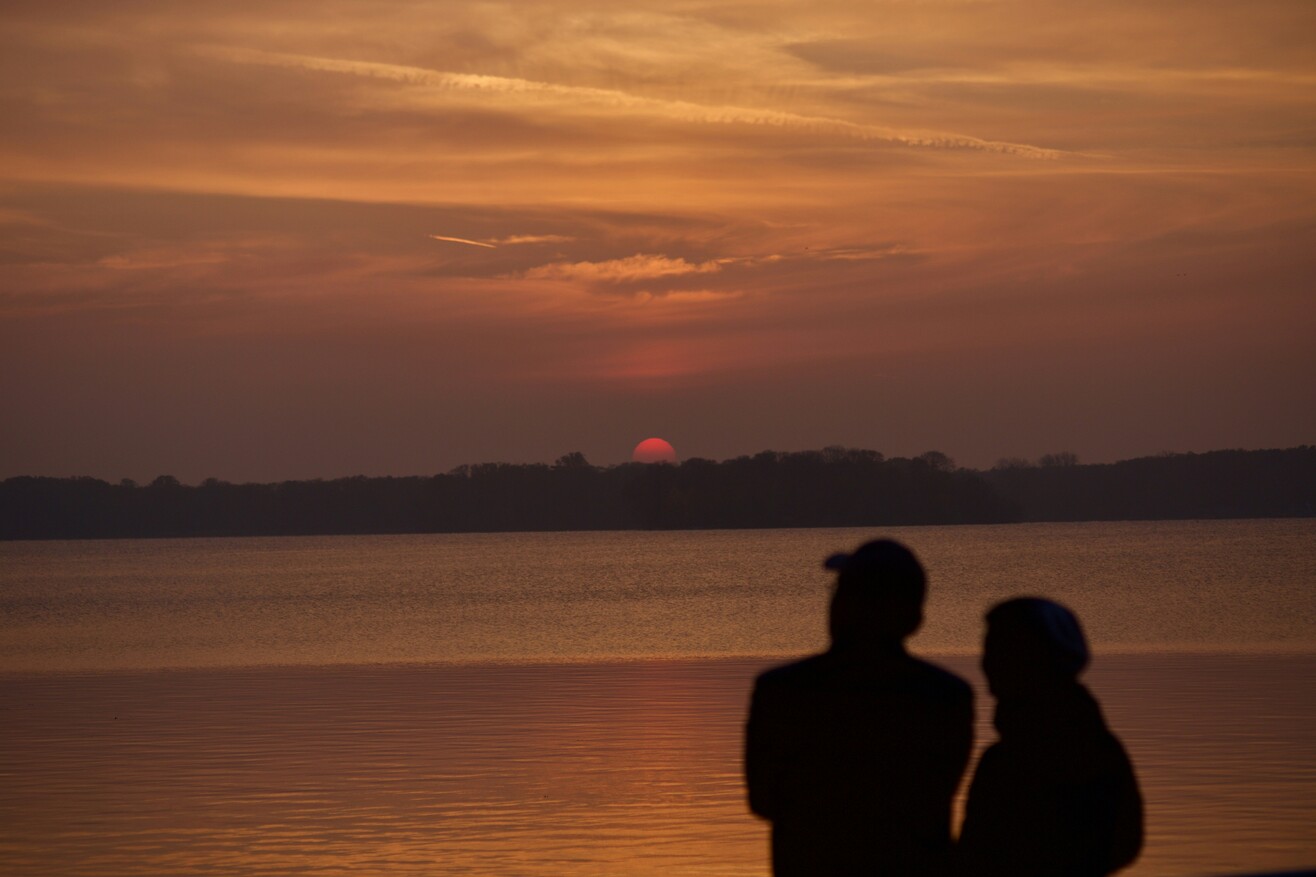 Zwei Personen stehen am Ufer des Tegeler Sees in Berlin und blicken bei Sonnenuntergang über das Wasser.