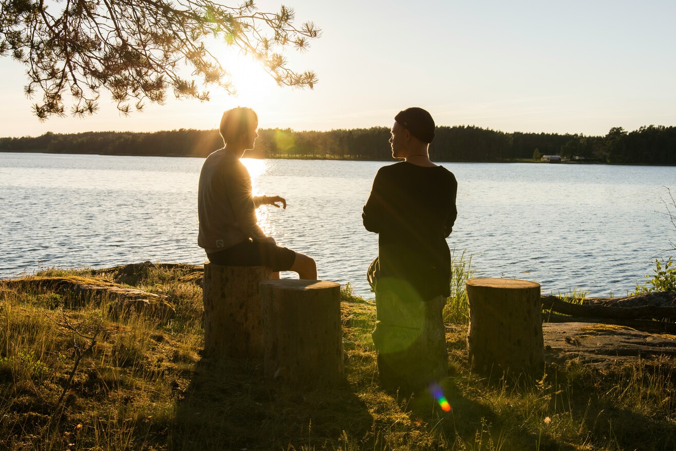 Zwei Personen stehen bei Sonnenuntergang an einem Gewässer und unterhalten sich miteinander.