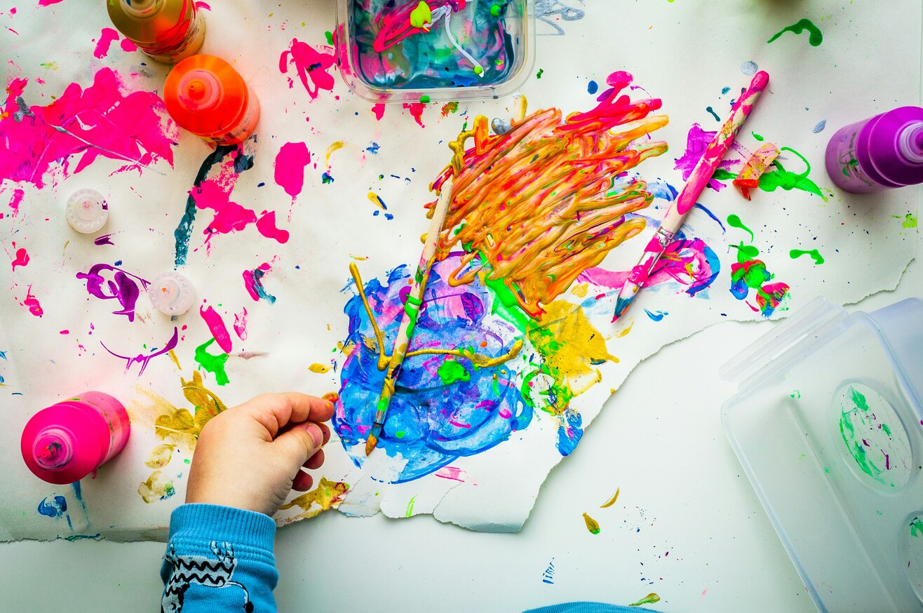 Bunte Gemälde und Malutensilien liegen auf einer Arbeitsfläche und zeigen verschiedene Formen kreativer Gestaltung.