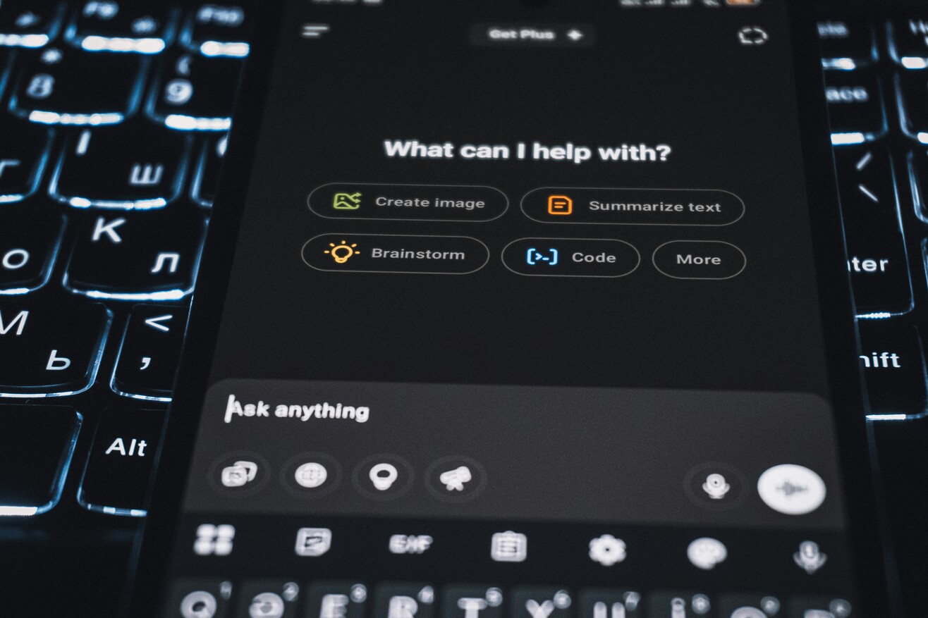 Smartphone in einer Hand, auf dessen Display die Oberfläche eines KI-Assistenten zu sehen ist.
